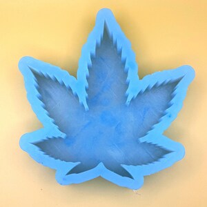 May include: A light blue silicone mold in the shape of a cannabis leaf. The mold has a detailed outline and a smooth interior. The background is a solid light yellow color.