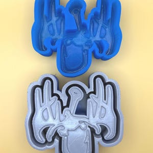 May include: Two cookie cutters, one blue and one gray, are displayed against a yellow background. The cutters are in the shape of a stylized design with the word "Hooked" and a fishing hook. The blue cutter is a solid form, while the gray one is an outline.