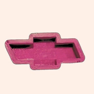 May include: A magenta-colored Chevrolet logo emblem. The emblem is a stylized bow tie shape with a rectangular outline and a recessed center. The emblem is set against a light beige background.