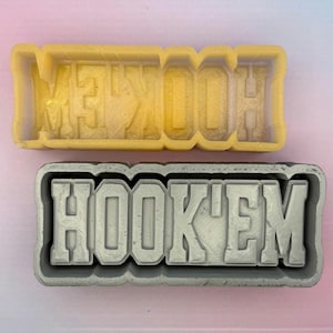 May include: Two rectangular molds, one yellow and one gray, each with the text "HOOK'EM" in bold, block letters. The yellow mold is empty, while the gray mold has the text raised.
