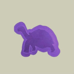 May include: A purple turtle-shaped object with a lighter purple outline, set against a light beige background. The turtle has a detailed form, with visible legs, head, and shell. The image is simple and clean.