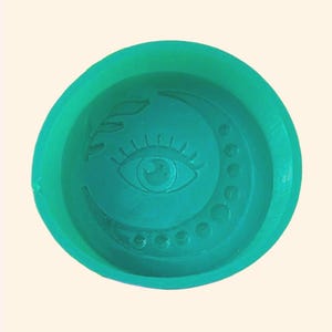 May include: A teal-colored, circular mold featuring an eye, crescent moon, and leaf design. The eye is centered, with the moon and dots to the right. The leaf design is to the left of the eye. The mold has a raised edge.