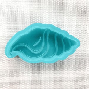 May include: A turquoise seashell-shaped mold. The mold is made of a flexible material and features detailed ridges and curves, resembling a natural seashell. The mold is designed for crafting and creating decorative items.