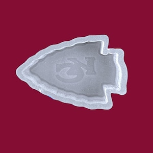 May include: A clear, arrowhead-shaped mold with the letters "KC" embossed in the center. The mold has a textured border and is set against a maroon background. This item is likely used for crafting.