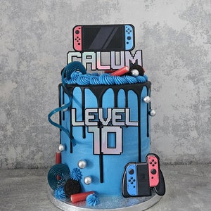 May include: A blue and black drip cake decorated with a Nintendo Switch controller, the name "CALUM" and "LEVEL 10" in pixel font. The cake is decorated with blue and black frosting, silver sprinkles, and red and blue candy.