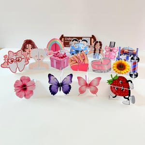May include: A collection of colorful, decorative paper cutouts, including butterflies, flowers, a gift box, a strawberry character, and other whimsical designs. The cutouts are arranged on a white surface.