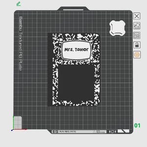 May include: A black and white composition notebook with the name "Mrs. Taylor" in a white label. The notebook is on a textured gray surface with a grid pattern. The image also shows a white object and some technical details.