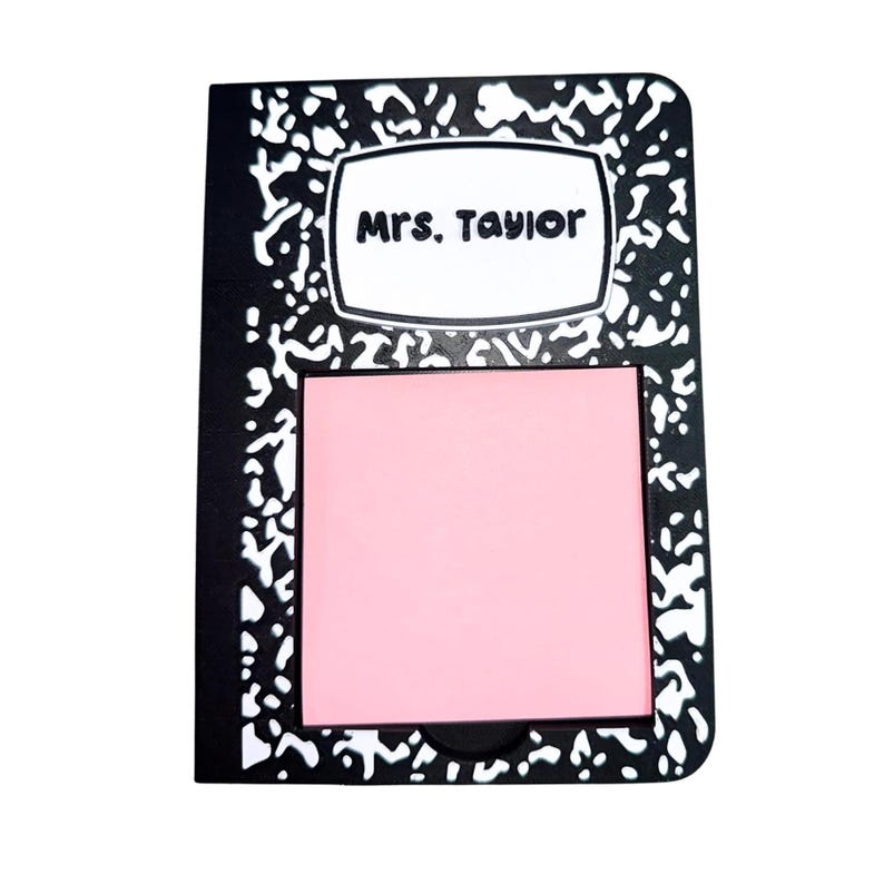 May include: A black and white notebook-style sticky note holder with the text "Mrs. Taylor" on a white label. A pink sticky note pad is in the center of the holder.