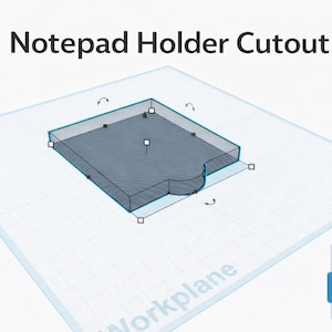 May include: A 3D model of a notepad holder cutout, rendered in a blue-gray color. The design includes a rectangular base with a curved cutout on one side. The text "Notepad Holder Cutout" is displayed above the model.