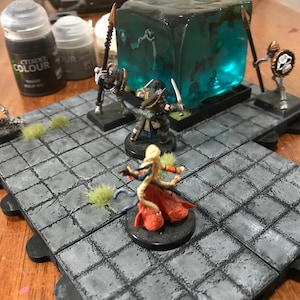 May include: A tabletop game scene with miniature figures, including a skeleton warrior, a human warrior, and a female warrior with a red and white dress. The figures are arranged on a gray and black tiled game board with green grass tufts. A clear blue cube with a skeletal figure inside is in the background.