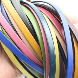 Könnte beinhalten: Eine Sammlung farbenfroher Lederriemen in verschiedenen Farbtönen, darunter Gelb, Rosa, Blau, Grün und Schwarz. Die Riemen sind eng aneinandergereiht und zeigen ihre glatte Textur und lebendigen Farben. Das Bild hebt die Vielfalt der verfügbaren Farben hervor.