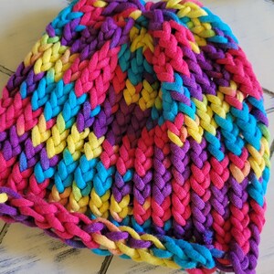 May include: A colorful knitted beanie hat with a braided pattern in shades of pink, purple, blue, and yellow.