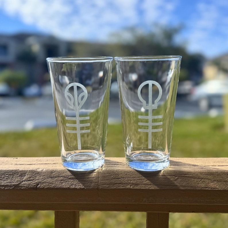 Puede incluir: Dos vasos de pinta de vidrio transparente con un dise&ntilde;o grabado de un s&iacute;mbolo blanco en cada uno. El s&iacute;mbolo es un c&iacute;rculo con una l&iacute;nea que lo atraviesa y un c&iacute;rculo m&aacute;s peque&ntilde;o en la parte superior.