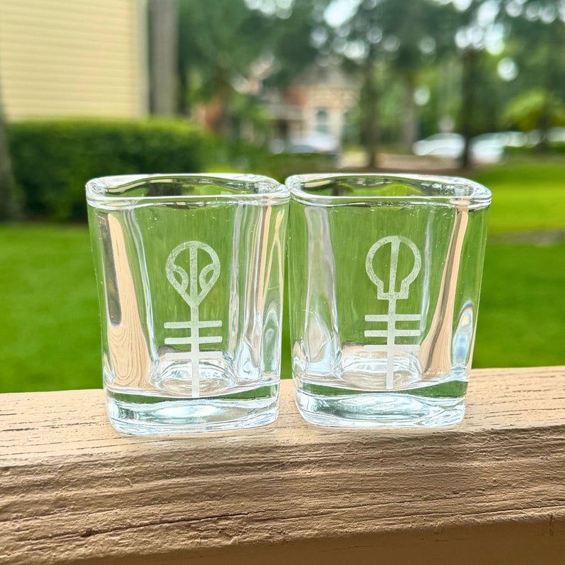 Puede incluir: Dos vasos de chupito de vidrio transparente con un logotipo blanco en la parte delantera. El logotipo es un s&iacute;mbolo estilizado con un c&iacute;rculo y l&iacute;neas.