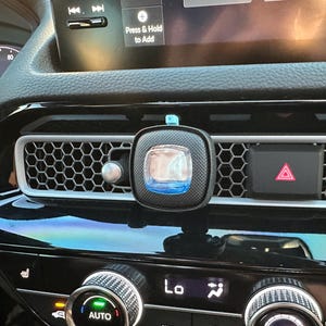 May include: A black car air freshener with a square frame and a blue translucent center, mounted on a honeycomb-patterned vehicle dashboard air vent next to a red hazard light button, with digital climate control dials visible below.