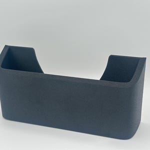 May include: A matte dark gray wall-mounted storage pocket with a rectangular base and rounded corners. The container features a wide, open top with a U-shaped cutout on the front panel, providing a clear view into the interior storage space.