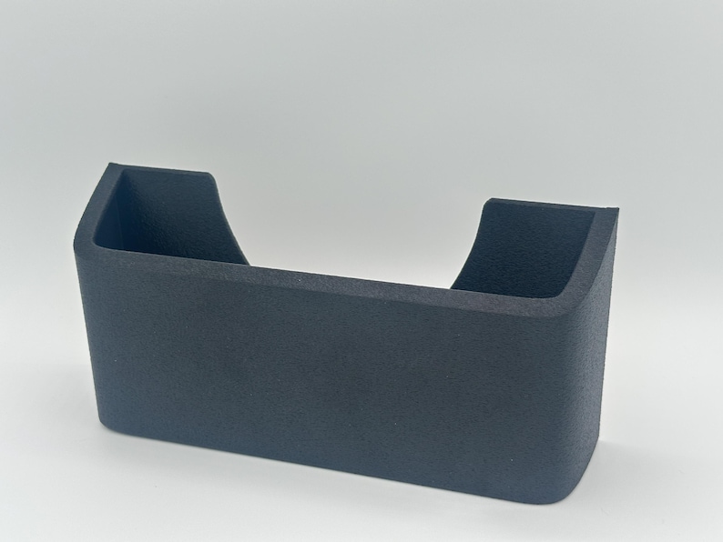 May include: A matte dark gray wall-mounted storage pocket with a rectangular base and rounded corners. The container features a wide, open top with a U-shaped cutout on the front panel, providing a clear view into the interior storage space.