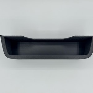 May include: A black, rectangular storage tray with a textured exterior finish and a smooth interior base. The tray features a slightly curved top edge along the front, creating a wide, open-top compartment designed for organizing small items.