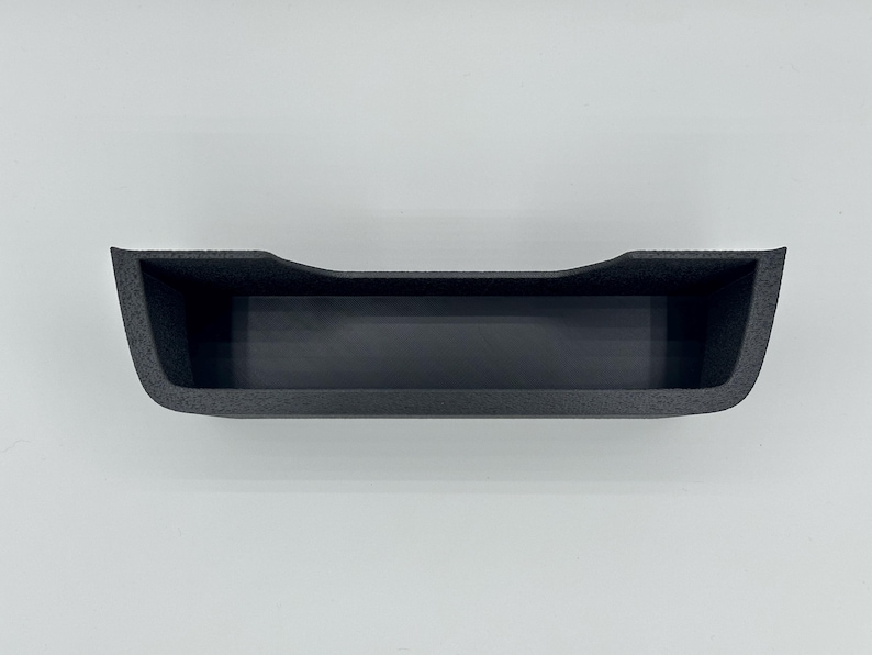 May include: A black, rectangular storage tray with a textured exterior finish and a smooth interior base. The tray features a slightly curved top edge along the front, creating a wide, open-top compartment designed for organizing small items.