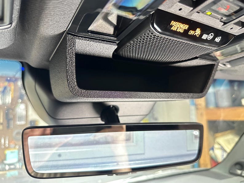May include: A black overhead console storage compartment mounted in a vehicle interior, positioned above the rearview mirror. The unit features a textured plastic finish and an integrated passenger airbag status indicator light with the text PASSENGER AIR BAG OFF.
