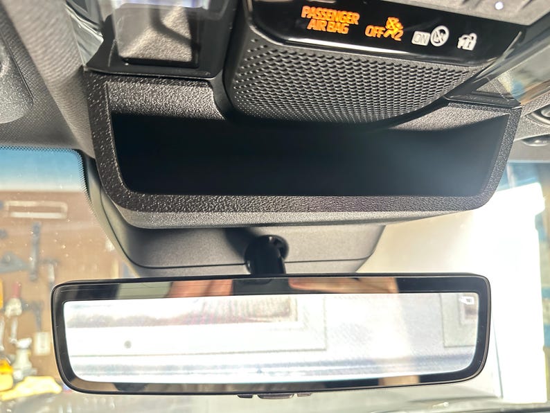 May include: A black overhead console in a vehicle featuring a storage compartment, a rearview mirror, and an illuminated display panel with the text PASSENGER AIR BAG OFF and various indicator icons for safety and vehicle status.
