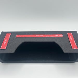 May include: A matte black rectangular storage bin with a wide front opening. The top surface features three strips of red 3M VHB adhesive tape, each printed with white text, designed for mounting the unit to a flat surface.