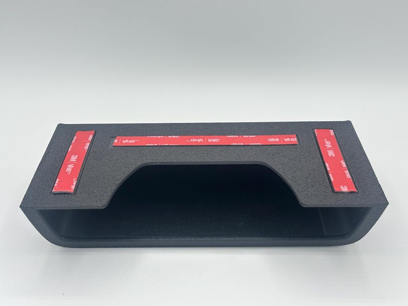 May include: A matte black rectangular storage bin with a wide front opening. The top surface features three strips of red 3M VHB adhesive tape, each printed with white text, designed for mounting the unit to a flat surface.