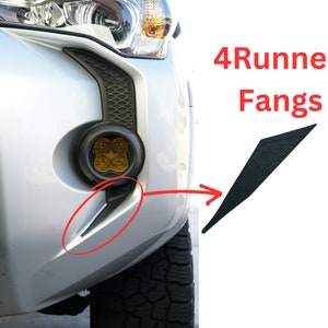 May include: A silver Toyota 4Runner with black 4Runner Fangs installed on the front bumper. The fangs are a pair of black, triangular-shaped pieces that extend from the bottom of the bumper.