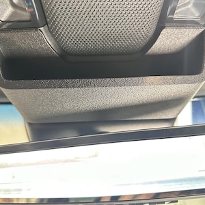 May include: A black plastic overhead console storage compartment with a textured finish, positioned directly above a rectangular rearview mirror. The storage unit features a deep, open bin design with a grid-patterned surface on the top section.