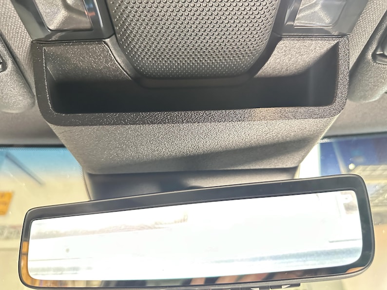 May include: A black plastic overhead console storage compartment with a textured finish, positioned directly above a rectangular rearview mirror. The storage unit features a deep, open bin design with a grid-patterned surface on the top section.