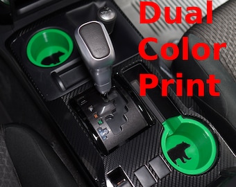 May include: Two black and green cup holders with a bear silhouette design are installed in a car's center console. The console has a carbon fiber pattern and includes a gear shifter and an automatic window switch. The text "Dual Color Print" is visible in the image.