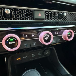May include: Three light pink textured covers fitted onto the circular climate control knobs of a vehicle dashboard. The knobs feature illuminated digital displays with text including AUTO, ON/OFF, and SYNC, set against a black glossy console.
