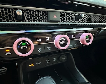May include: Three light pink textured covers fitted onto the circular climate control knobs of a vehicle dashboard. The knobs feature illuminated digital displays with text including AUTO, ON/OFF, and SYNC, set against a black glossy console.