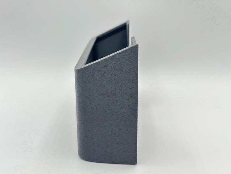 May include: A dark gray, U-shaped wall-mounted holder with a textured, matte finish. The object features a rectangular front panel, two side walls, and an open top, designed with a clean, geometric structure and a solid, uniform appearance.