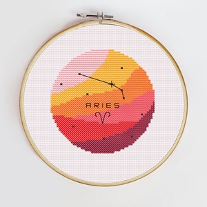 Può includere: Un cerchio da ricamo a punto croce con uno schema di colori rosa, arancio e rosso. Il disegno presenta la costellazione zodiacale dell'Ariete, con la parola "ARIES" e il simbolo dell'Ariete al centro.