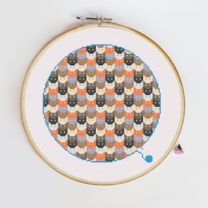 Puede incluir: Un patrón circular de punto de cruz con un diseño repetitivo de caras de gato estilizadas en varios colores, incluyendo naranja, negro, gris y blanco. El patrón está enmarcado en un aro de bordado de madera.