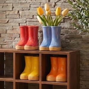 May include: A wooden shelf unit displays colorful rain boot vases. Red, blue, yellow, and orange boots hold yellow tulips. The background features a stone wall and a plant. The text "Malinda Boutiques" is in the upper left corner.