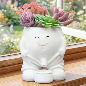 May include: A white ceramic planter shaped like a person, with a smiling face and a small pot at the base. The planter is filled with a variety of succulents in shades of green, pink, and purple. The planter is sitting on a wooden surface.