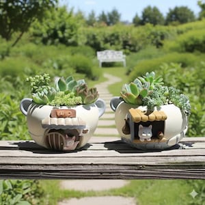 May include: Two cream-colored ceramic planters, each shaped like a mug with a handle, house succulents. One has a cafe sign and a cat peeking from a doorway. The other has a cat in a dog house. The planters sit on a wooden surface.