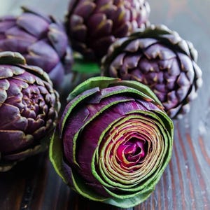 May include: Close-up of several purple artichokes with green-edged leaves. The artichokes are arranged on a dark wooden surface. The center of one artichoke is visible, revealing a spiral pattern.