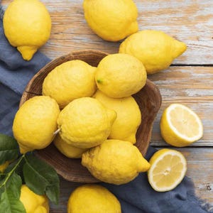 May include: A wooden bowl filled with bright yellow lemons, some whole and some sliced in half, sits on a rustic wooden surface. The lemons are surrounded by a blue cloth and green leaves, creating a fresh, vibrant display.