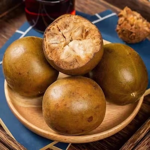 May include: A wooden plate holds four brown, round fruits, one cut in half to reveal a light-colored interior. The fruits have a smooth, slightly textured surface. The background includes a blue patterned cloth and a glass of red liquid.