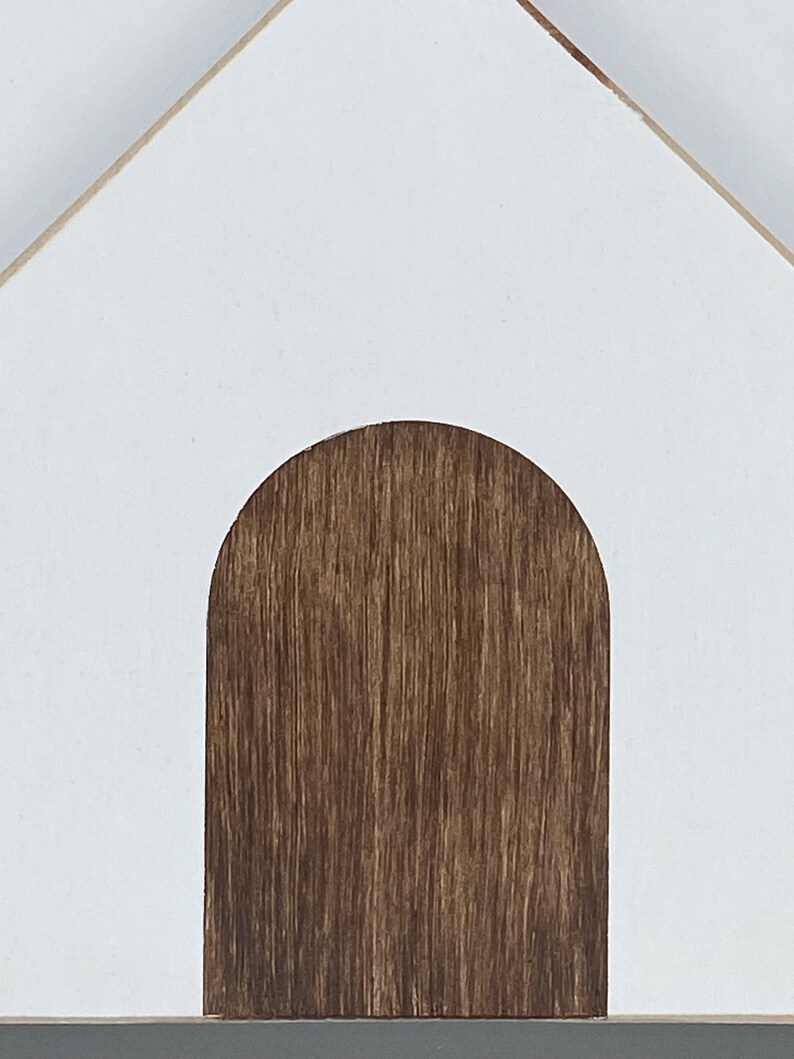 May include: White wooden house with a brown wooden door. The house is a simple design with a rounded archway for the door.