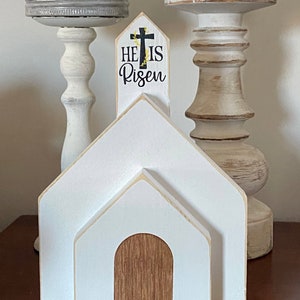 May include: A white wooden church with a brown wooden door and the text "He is Risen" on the front. The church is 7.75 inches tall, 5.5 inches wide, and 2.25 inches deep.
