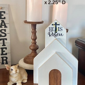 May include: White wooden church with a brown wooden door and a sign that reads "He is Risen" with a cross and floral accents. The church is 10.75 inches tall, 7.25 inches wide, and 2.25 inches deep.