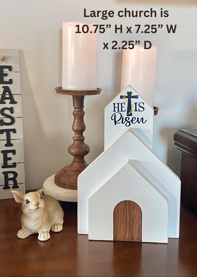 May include: White wooden church with a brown wooden door and a sign that reads "He is Risen" with a cross and floral accents. The church is 10.75 inches tall, 7.25 inches wide, and 2.25 inches deep.