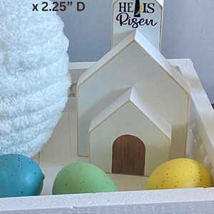 May include: White wooden church with a steeple that reads "He Is Risen". The church is sitting in a white wooden tray with a decorative trim. There are three speckled Easter eggs in the tray, one green, one blue, and one yellow.