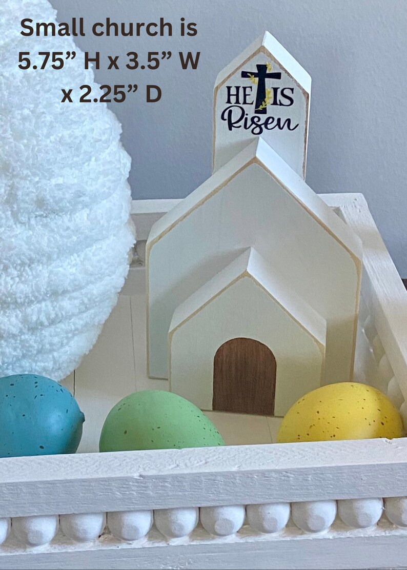 May include: White wooden church with a steeple that reads "He Is Risen". The church is sitting in a white wooden tray with a decorative trim. There are three speckled Easter eggs in the tray, one green, one blue, and one yellow.