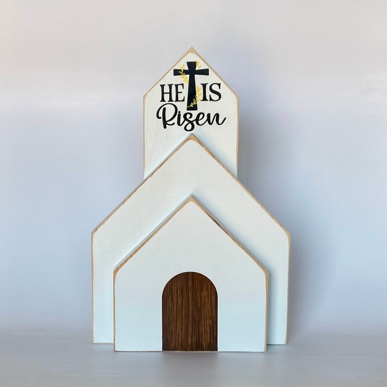 May include: White wooden church-shaped tiered decor with a black cross and the text "He is Risen" on the top tier.