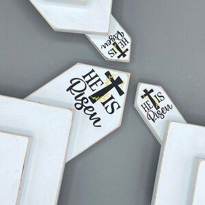 May include: Three white wooden signs with a chevron shape. Each sign has the text "He is Risen" with a black cross and yellow floral accents.